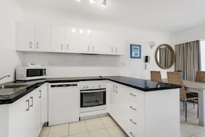 A modern kitchen with white cabinets, a black countertop, a microwave, a dishwasher, and an oven. There is a dining area with wicker chairs and a round mirror on the wall.