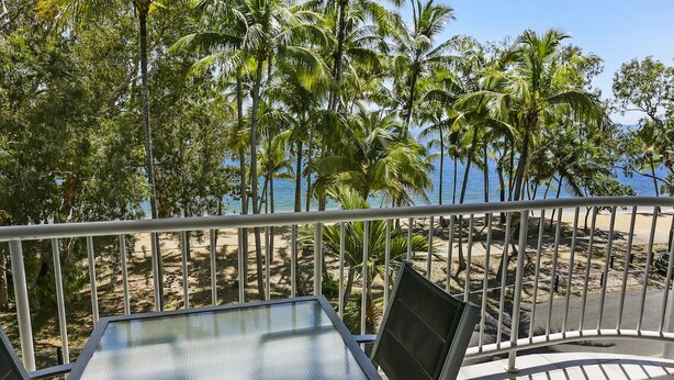 Un balcon avec une table et des chaises en verre, donnant sur une plage bordée de palmiers et sur l’océan.