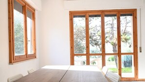 A room with a wooden table, white chairs, large windows, and a view of trees and a fence. - S077 - Sirolo, three-room apartment with garden in the center (Sirolo)