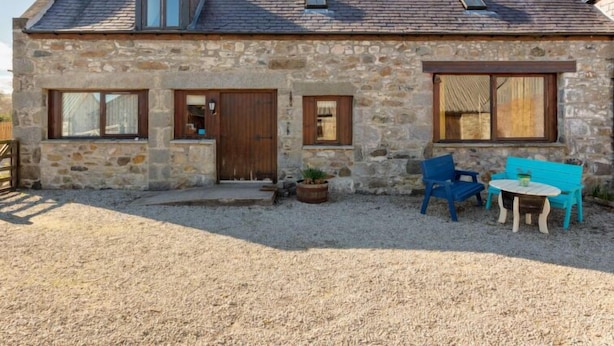 Una casa de piedra con techo inclinado, una puerta de madera y un pequeño porche con una mesa y sillas.