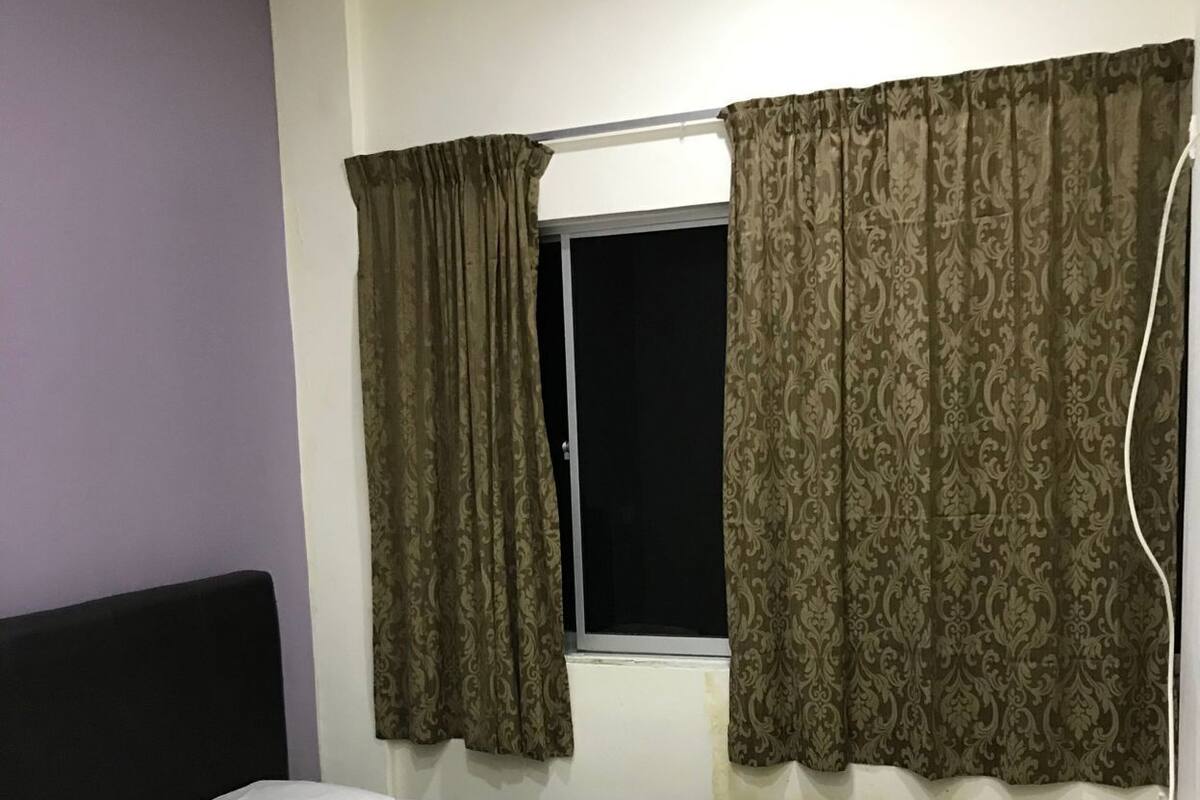 A single bed with a white mattress and a brown blanket, a window with patterned curtains, and a plain wall.