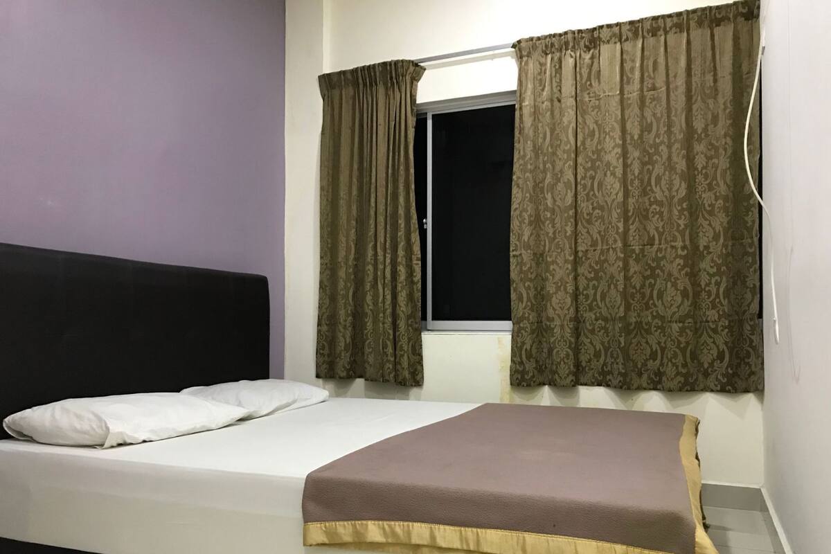 A single bed with a headboard, a window with curtains, and a wall with a power outlet.