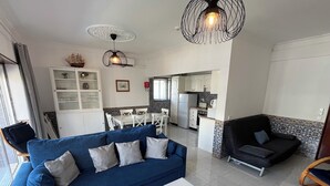 Un salón con un sofá azul, una mesa de centro blanca y un sillón negro. Se ve una cocina al fondo con armarios blancos y un salpicadero de azulejos.
