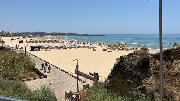 Una playa con un paseo marítimo de madera, sombrillas y un faro a lo lejos.