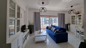 Una sala moderna con un sofá azul, una mesa de centro blanca y un televisor. Se observan dos lámparas colgantes, un armario con puertas de vidrio y un balcón a través de la ventana.