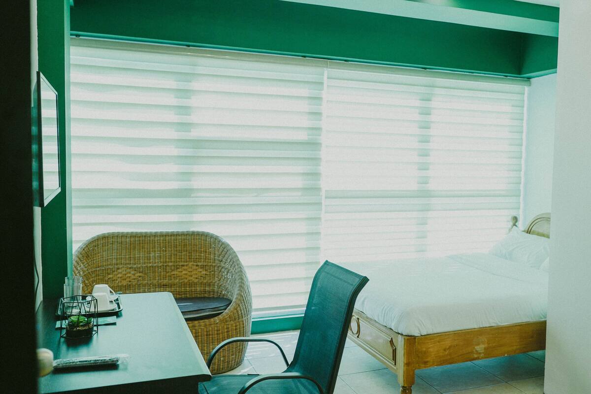 A room with a bed, a wicker chair, a table with a glass, and a window with blinds.
