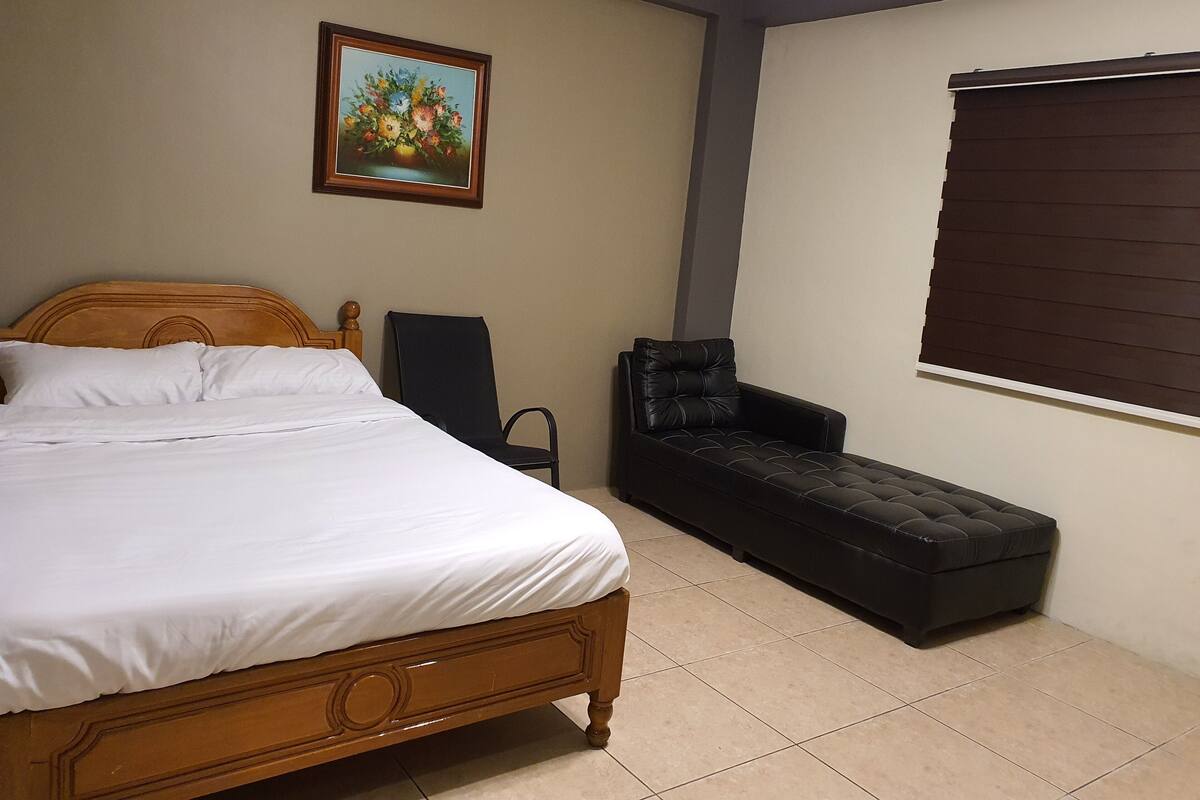 A bedroom with a wooden bed, a black armchair, a small sofa, a painting on the wall, and a window with blinds.