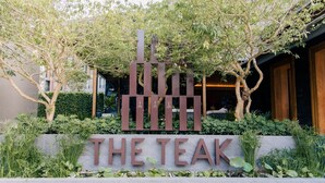 A sign with the name "THE TEAK" in large letters, surrounded by greenery and trees. - The Teak Hotel (Mae Sot)