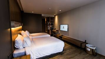 A modern hotel room with two beds, a wooden bench, a desk, a TV, and a trash bin.