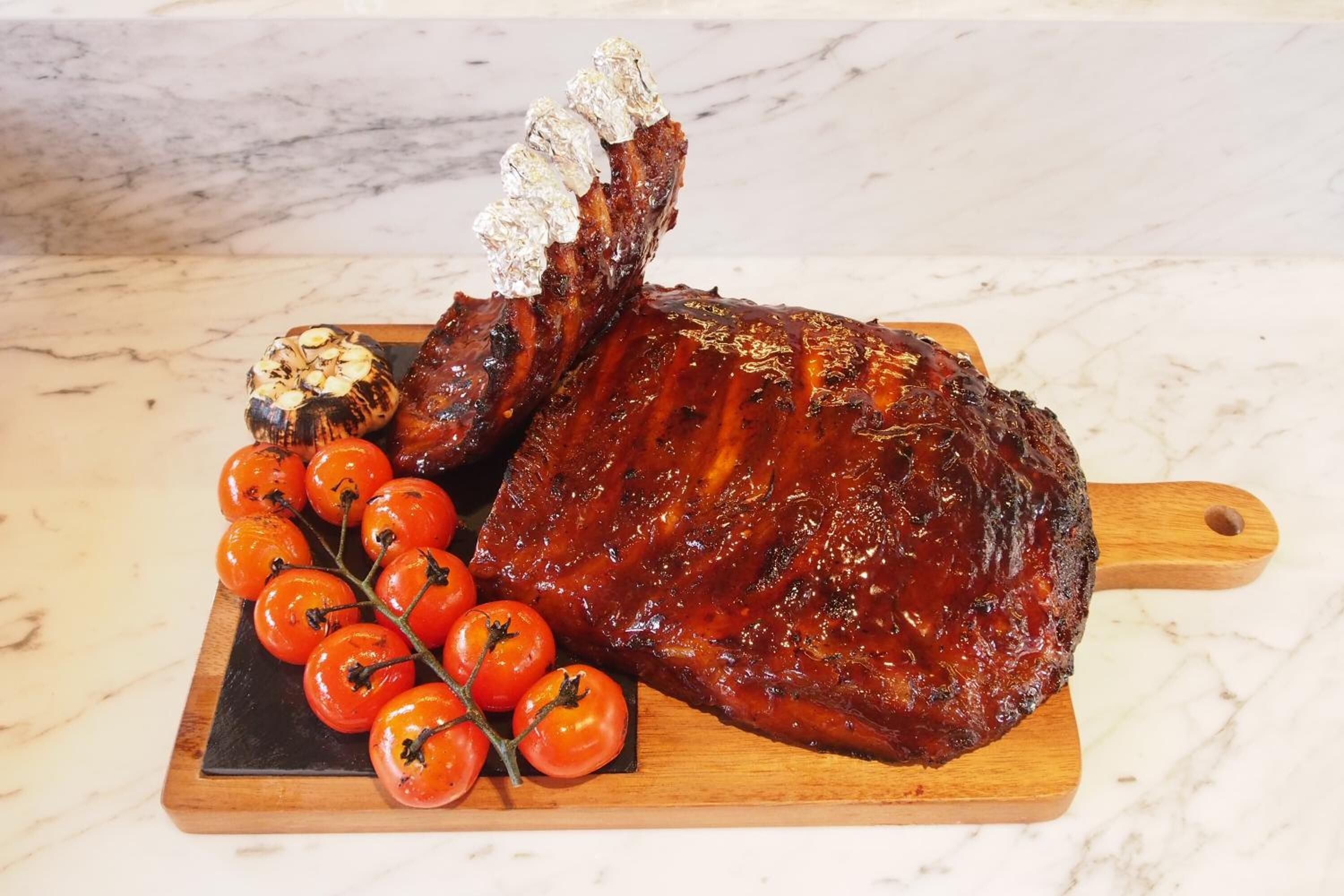 Un jarret de porc rôti à la peau croustillante, servi sur une planche en bois avec des tomates cerises et une tête d’ail.