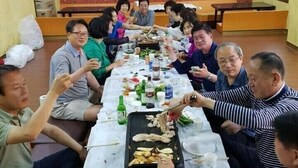 Un grupo de personas sentadas alrededor de una mesa larga con comida y bebidas, en una habitación con pared estampada y una ventana.