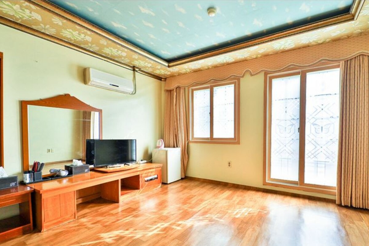 Una habitación con un escritorio de madera, televisión y un amplio ventanal con cortinas.