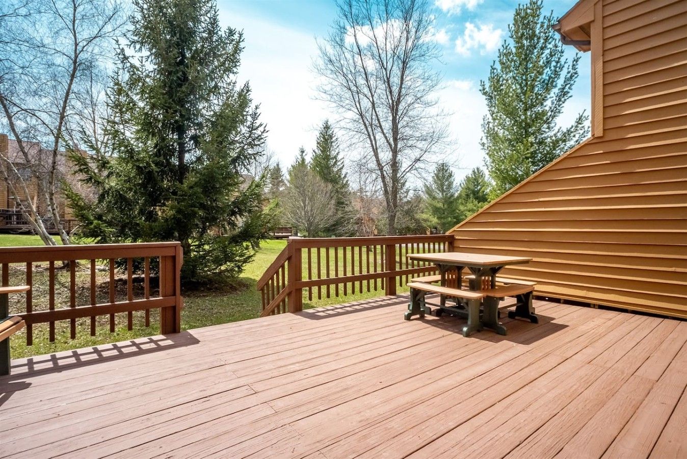 A wooden deck with a picnic table and benches, surrounded by trees and a building.