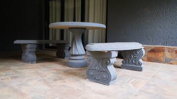 Un espace patio avec trois tables et chaises en pierre, un sol carrelé et un mur sombre agrémenté d’une fenêtre et de rideaux en arrière-plan.