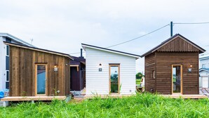 Trois petites cabanes en bois, chacune ayant un style distinctif et comprenant une porte et une petite fenêtre, sont installées dans une zone herbeuse sous un ciel dégagé.