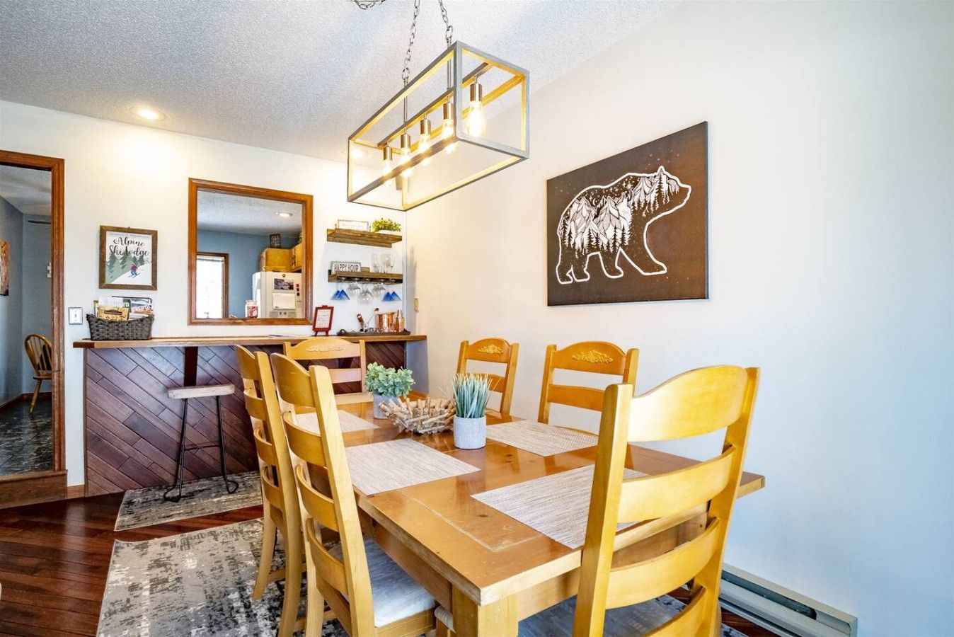 A dining area with a wooden table, chairs, a hanging light fixture, and a bear-themed artwork on the wall.