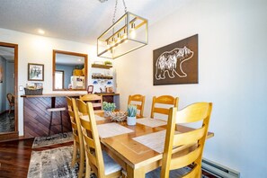 Un comedor con mesa de madera, sillas, una lámpara colgante y una obra de arte con motivos de oso en la pared.