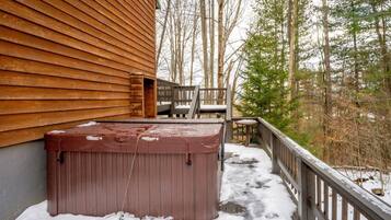 A wooden deck with a hot tub and a chair, surrounded by snow and trees.
