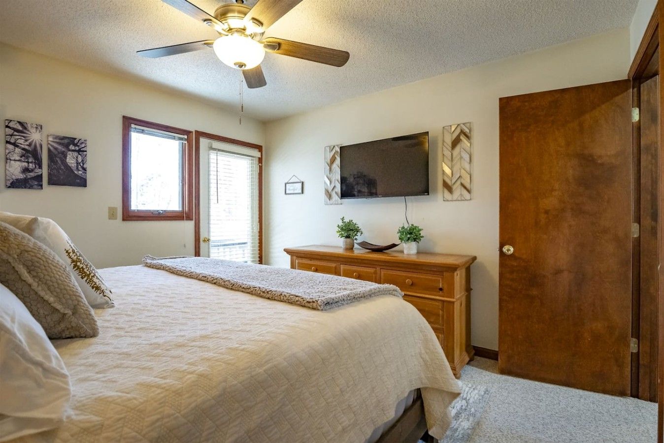 A bedroom with a bed, a ceiling fan, a TV, a wooden dresser, and a door.