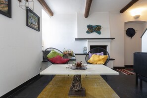 Een eethoek met een witte tafel, twee zwarte stoelen met kleurrijke kussens, een rustieke houten onderkast en een decoratieve plant als middelpunt.