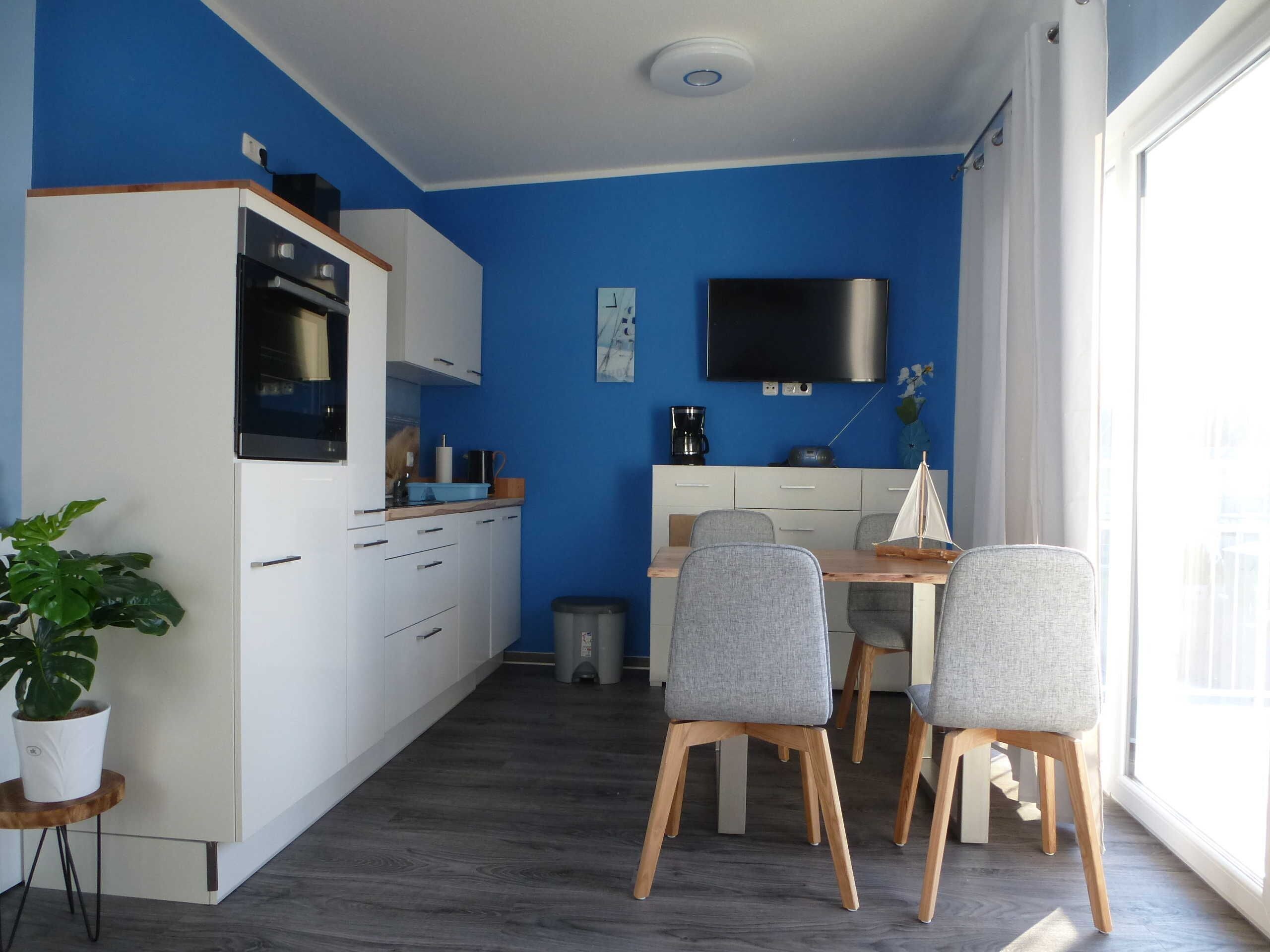 Een kleine keuken met witte kastjes, een magnetron en een koffiezetapparaat. Er is een eethoek met een houten tafel en grijze stoelen.