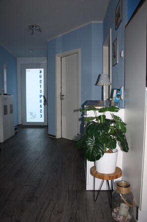 Um corredor com um vaso de planta, uma luminária e um pote de vidro sobre uma mesinha.