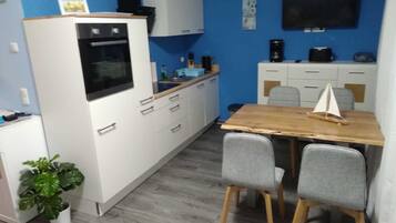 Een kleine keuken met witte kastjes, een houten tafel, twee grijze stoelen, een kleine potplant en een wandtelevisie.