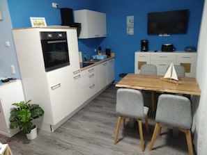 Une cuisine compacte avec des meubles blancs, une table en bois, deux chaises grises, une petite plante en pot et un téléviseur fixé au mur.