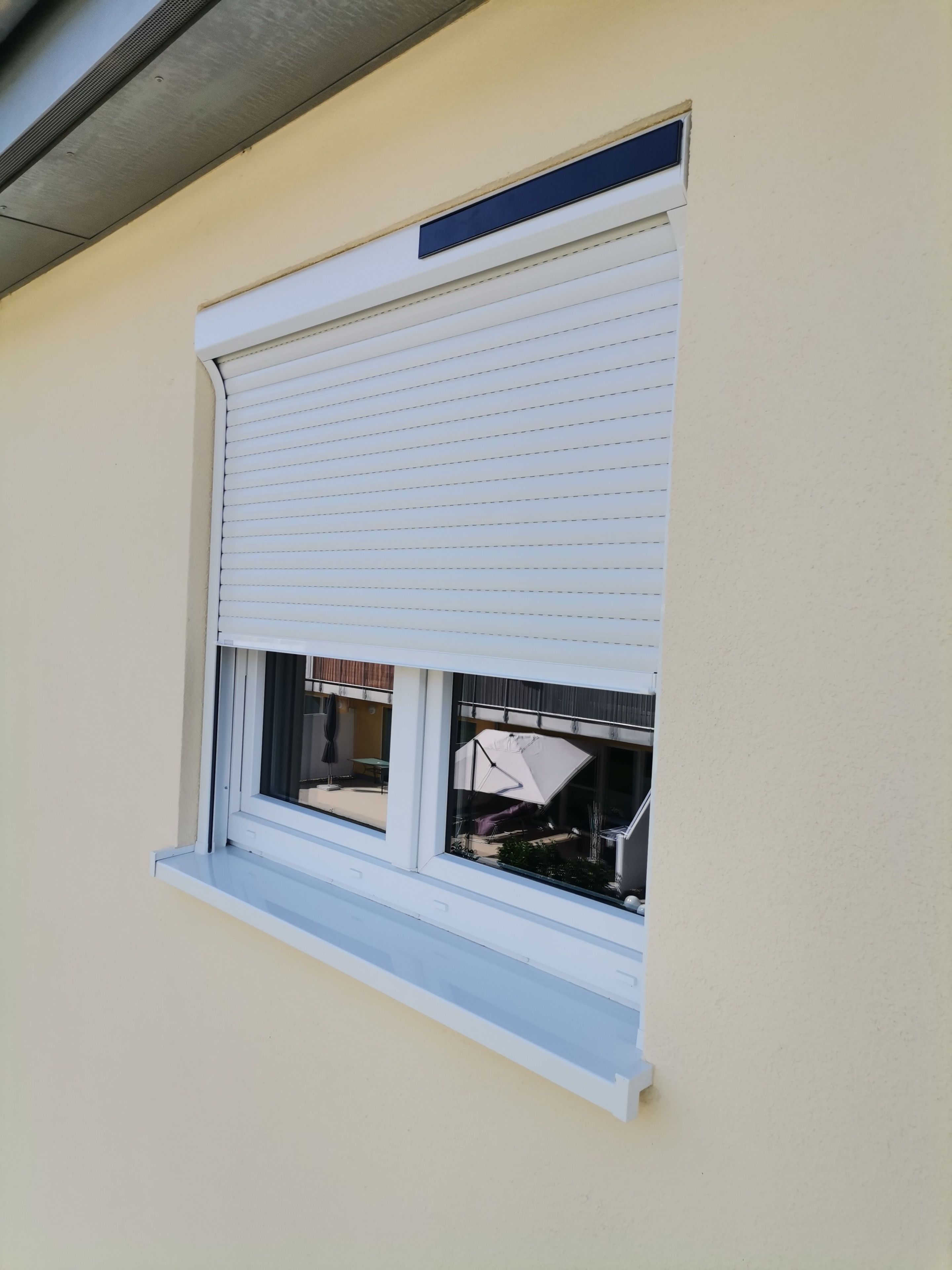 Ein Fenster mit weißem Rollo, weißem Fensterrahmen und einer kleinen Lüftung darüber.