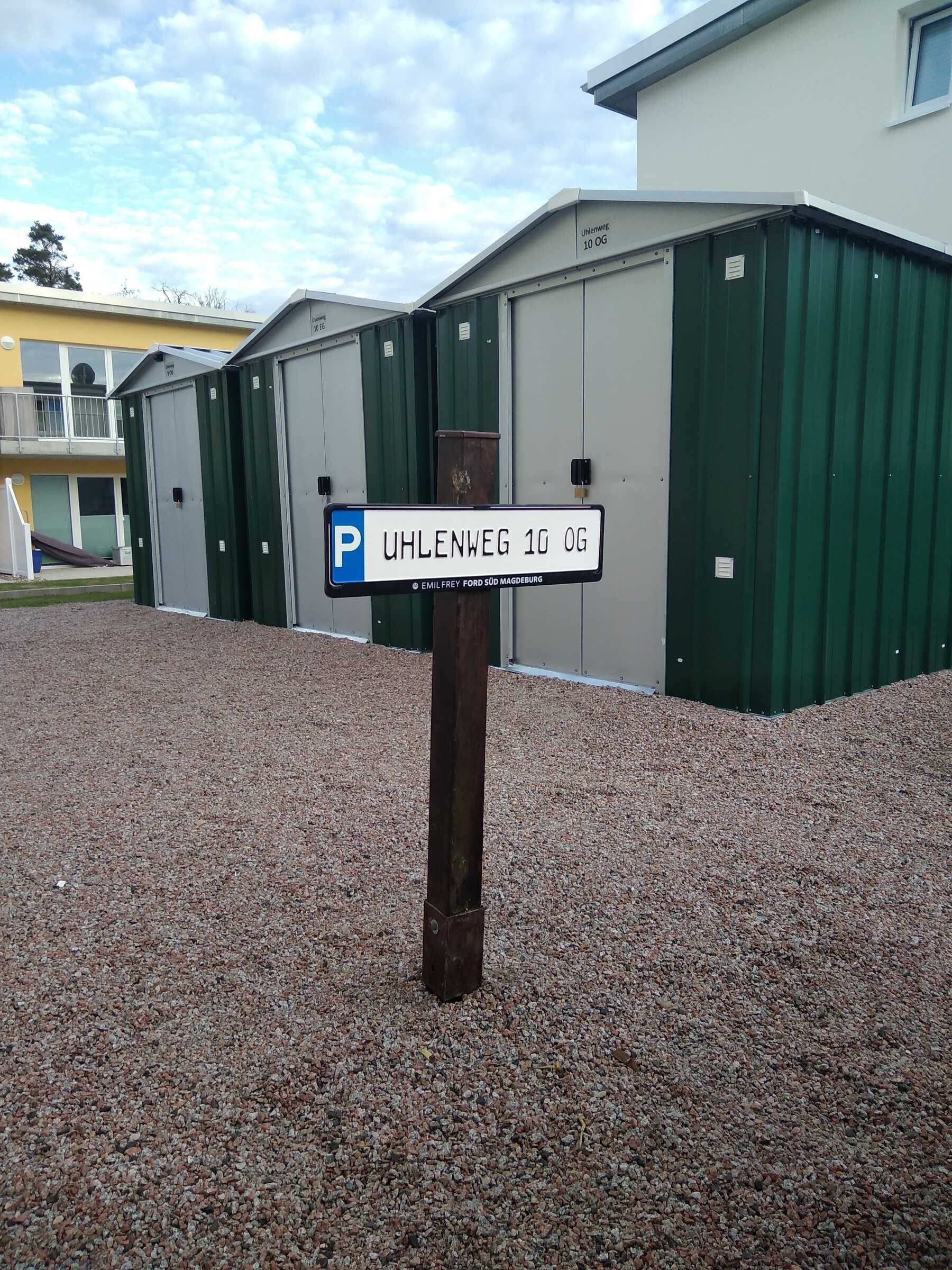 A parking sign for Uhlenweg 10 OG, a building with green and white exterior walls, and a gravel ground.