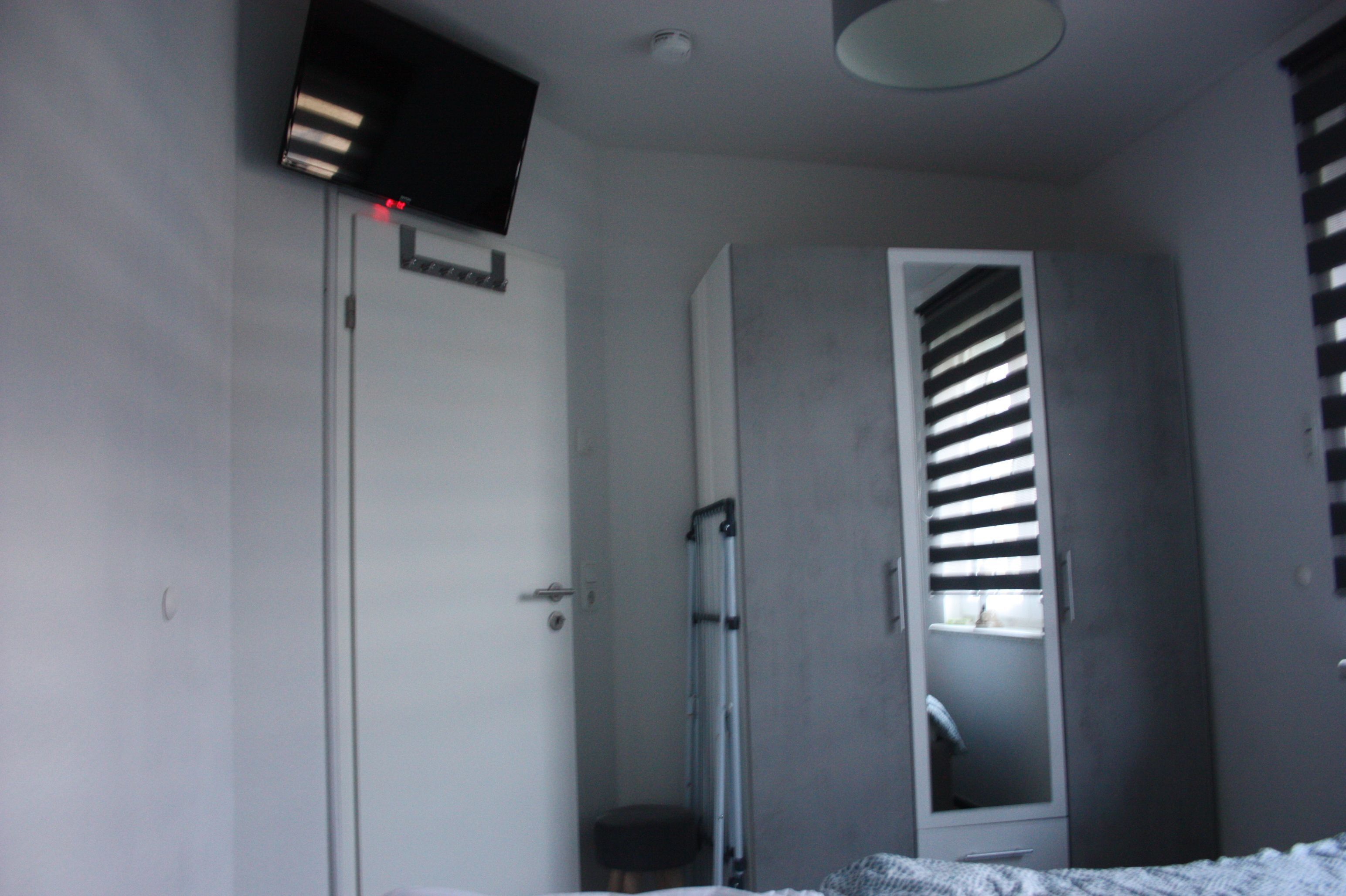 Ein Schlafzimmer mit einem Bett, einer Tür, einem Fenster mit Jalousien, einer Deckenleuchte und einem an der Wand montierten Fernseher.
