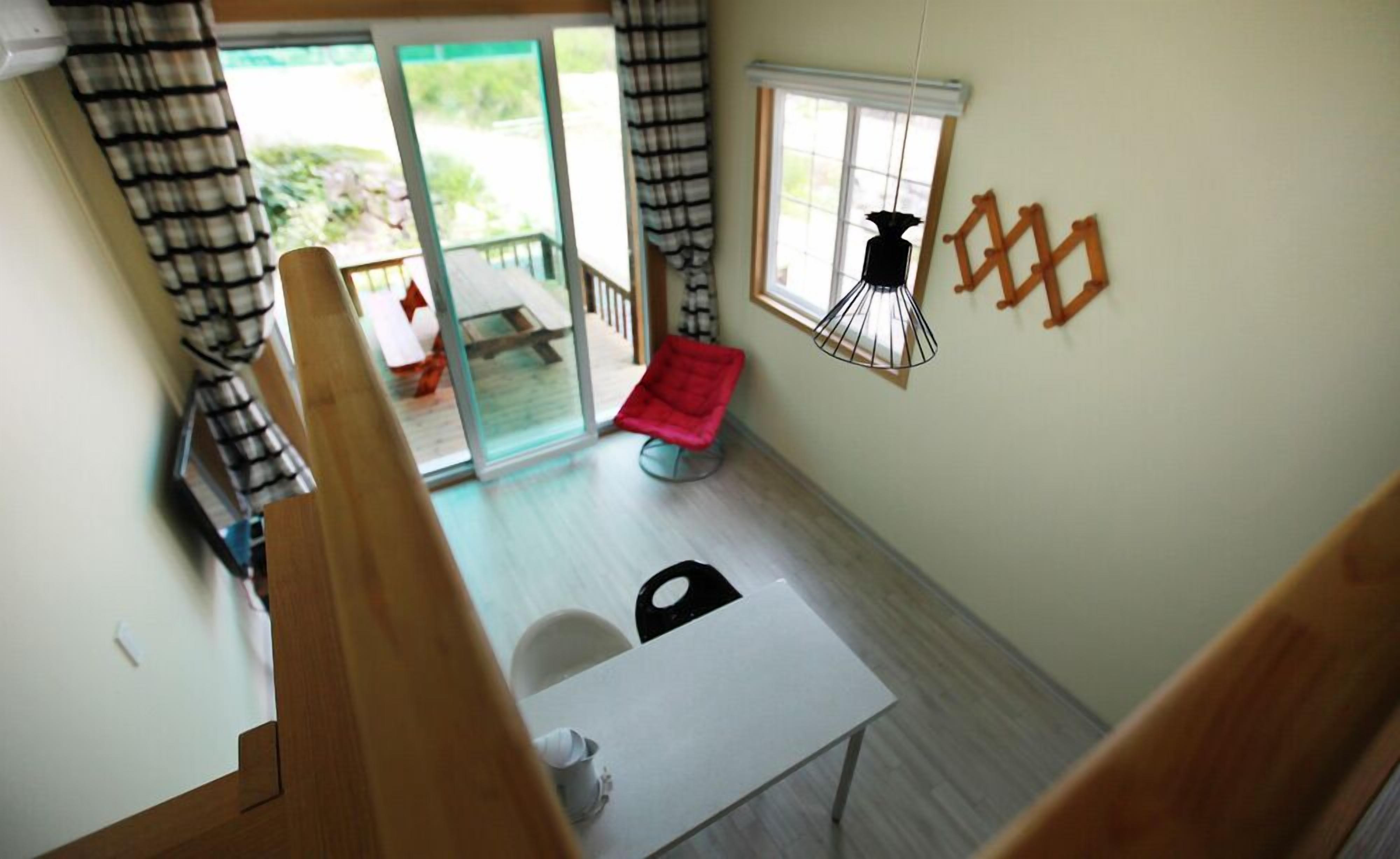 A room with a white table, a red chair, a window with a hanging decoration, and a view of a balcony with a red chair.