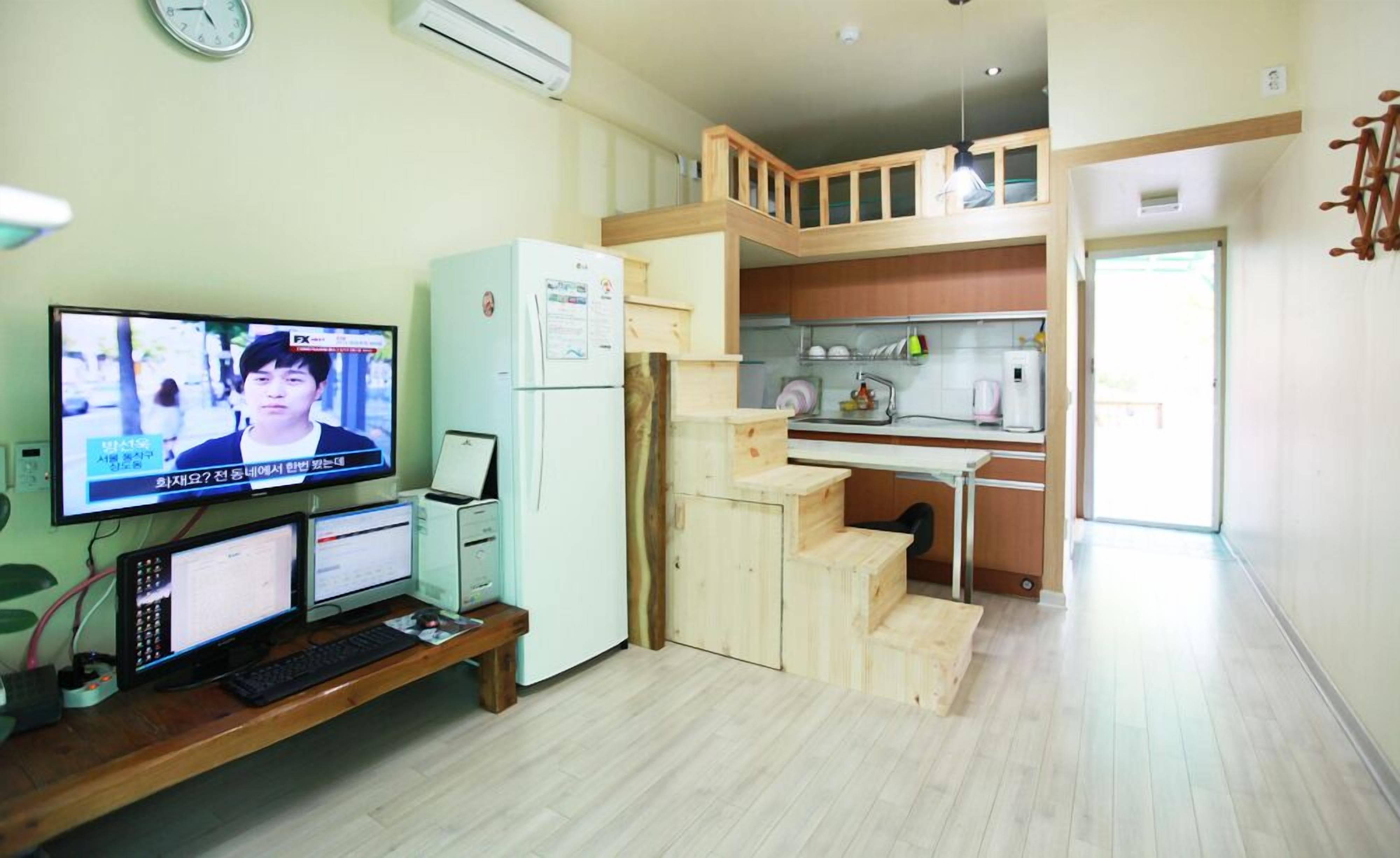 A compact living space with a kitchenette, a dining area, and a TV displaying a news program.