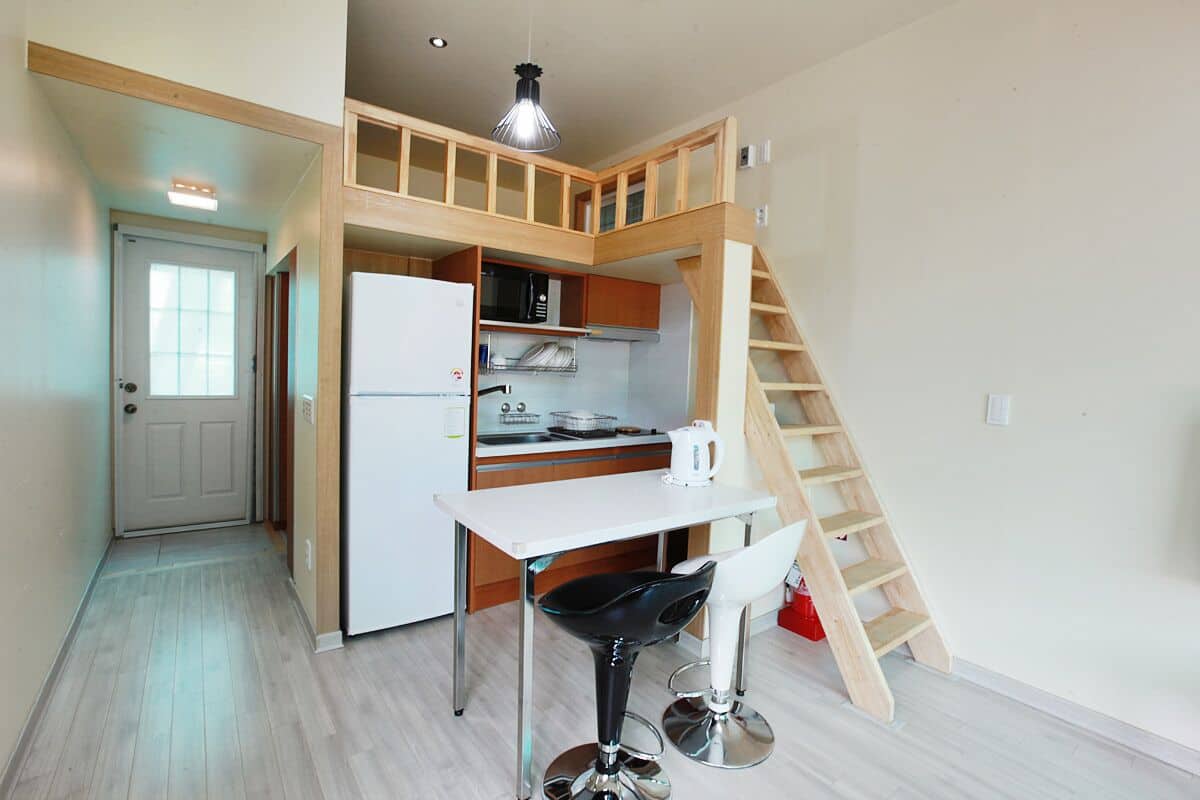 A compact kitchen with a white refrigerator, wooden cabinets, a white table, and a black chair. A wooden staircase leads to an upper level.