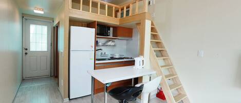 Una cocina compacta con un refrigerador blanco, armarios de madera, una mesa blanca y una silla negra. Una escalera de madera conduce a un nivel superior.