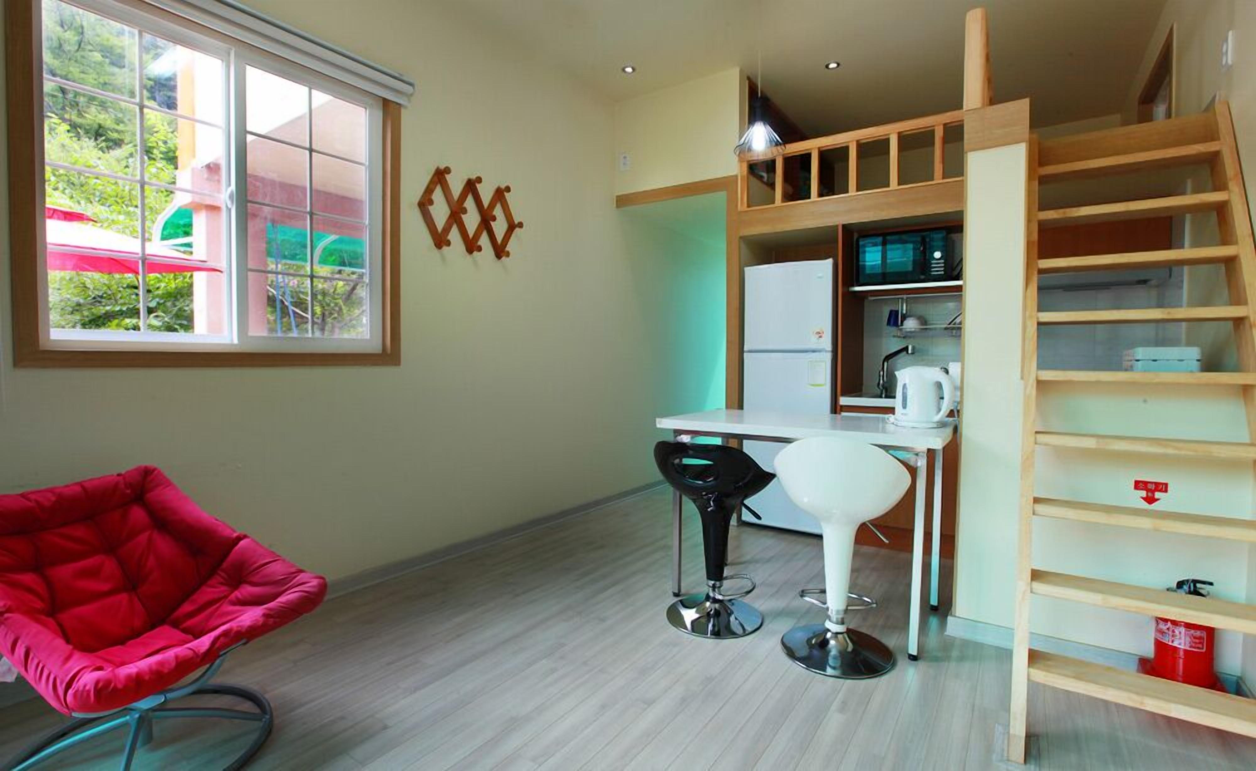 A compact room with a desk, two chairs, a small kitchenette, and a window with a view of greenery.