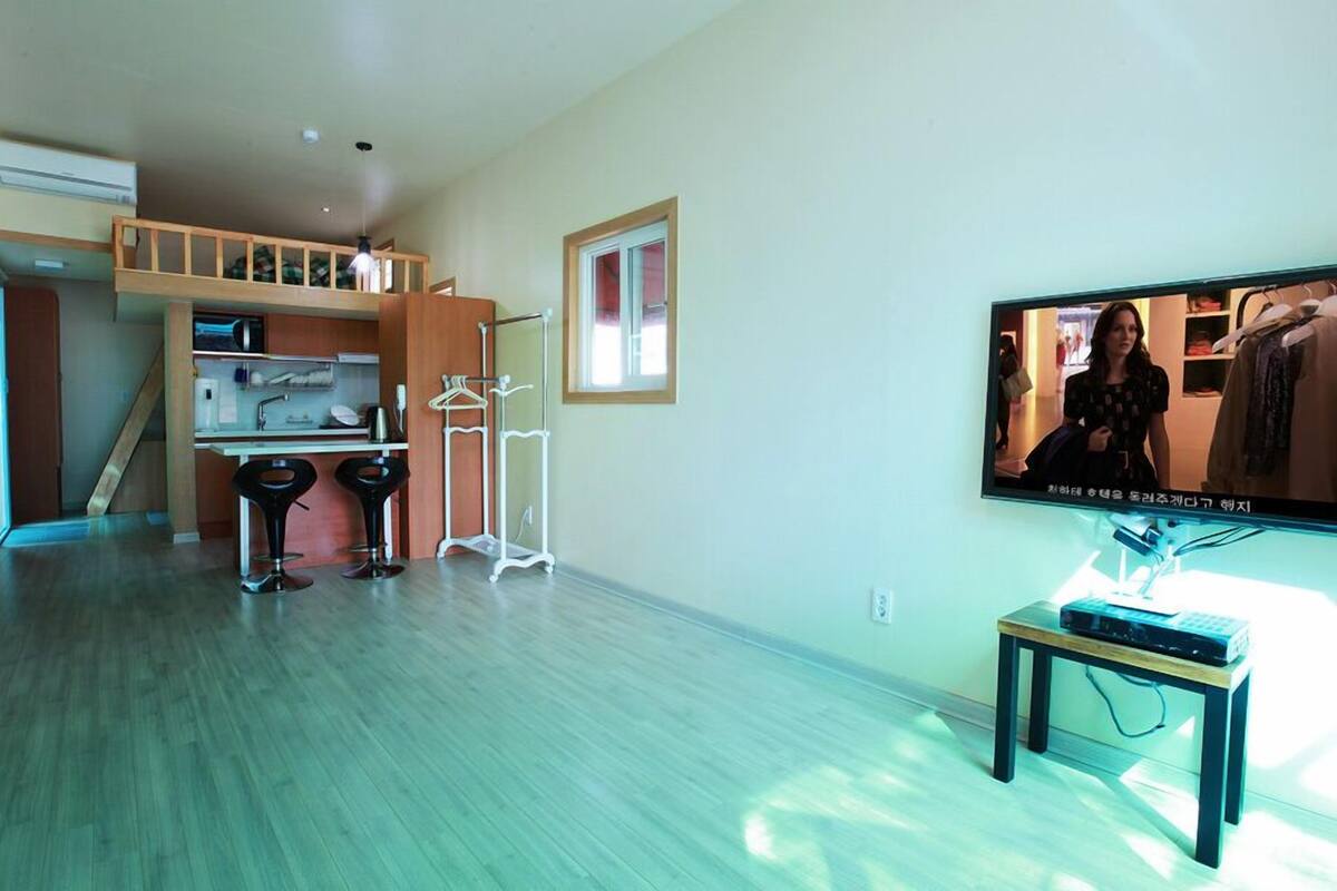 A modern living room with a kitchen area, a TV mounted on the wall, and a small table with electronic devices.
