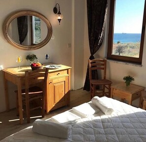 A bedroom with a bed, a wooden desk, a mirror, a small table, and a view of the sea. - Sukha Inn Otel & Beach (Bozcaada)