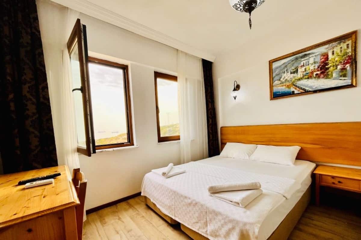 A bedroom with a wooden bed, two windows, a painting on the wall, and a wooden desk with a remote control.