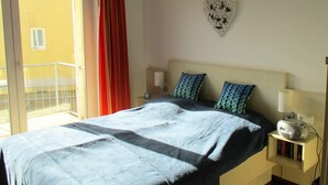 Een slaapkamer met een bed, twee kussens, een nachtkastje met een lamp, een klein plankje met boeken, een hartvormige decoratie aan de muur en een schuifdeur naar een balkon.