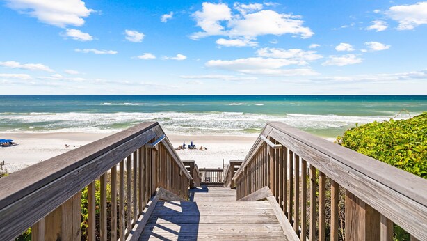 Una escalera de madera que lleva a una playa, con cielo azul despejado y olas del océano.