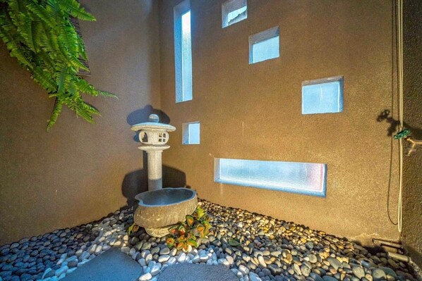 A stone water feature with a tiered design, surrounded by pebbles and small plants, against a textured wall with rectangular cutouts.