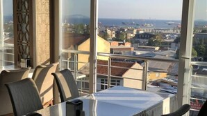 Una habitación de hotel moderna con un amplio ventanal que ofrece vistas a la ciudad, una mesa de comedor con sillas y una mesita con tetera y tazas.