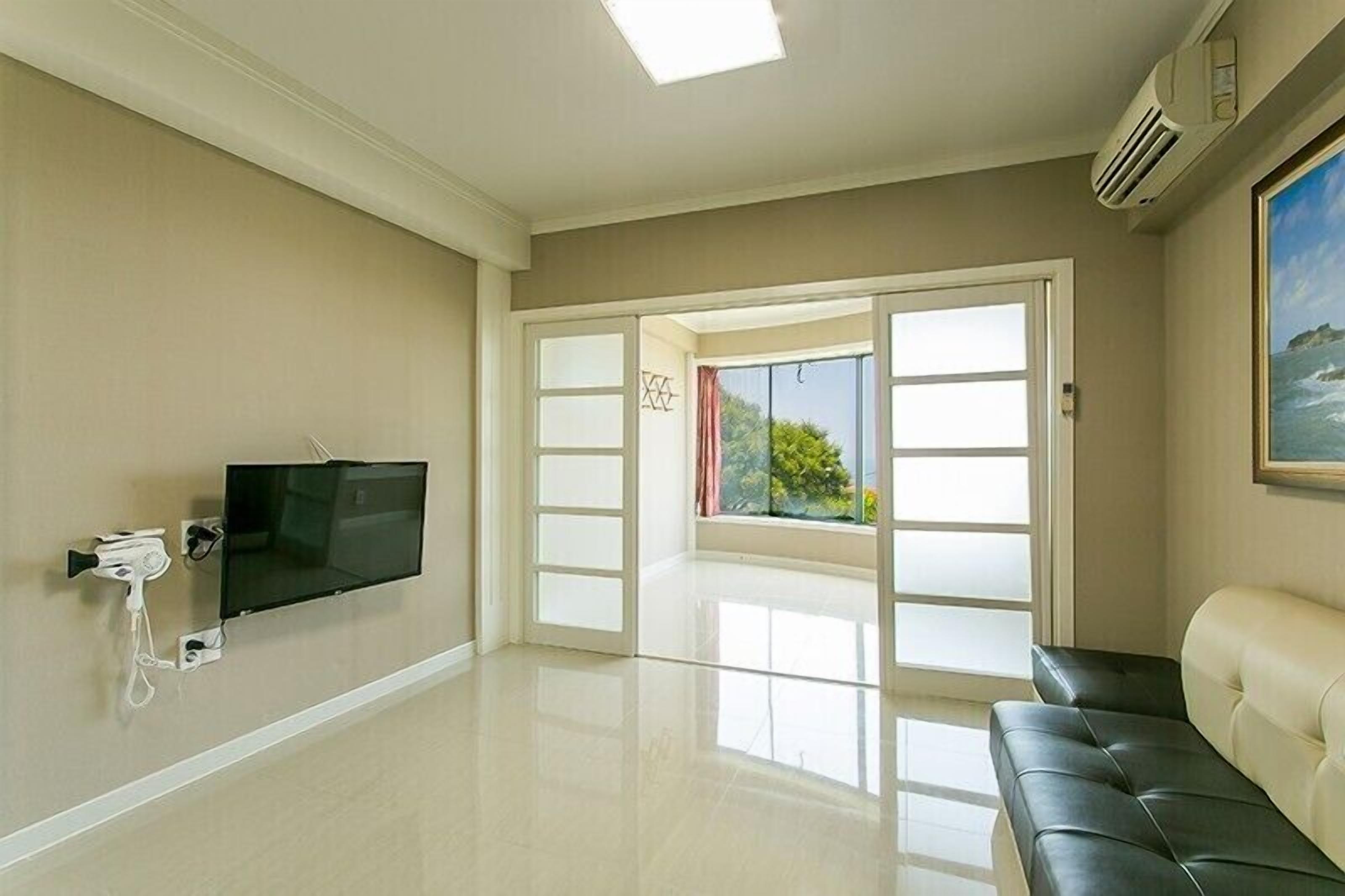 A spacious room with a large window, a flat-screen TV mounted on the wall, and a black leather sofa.