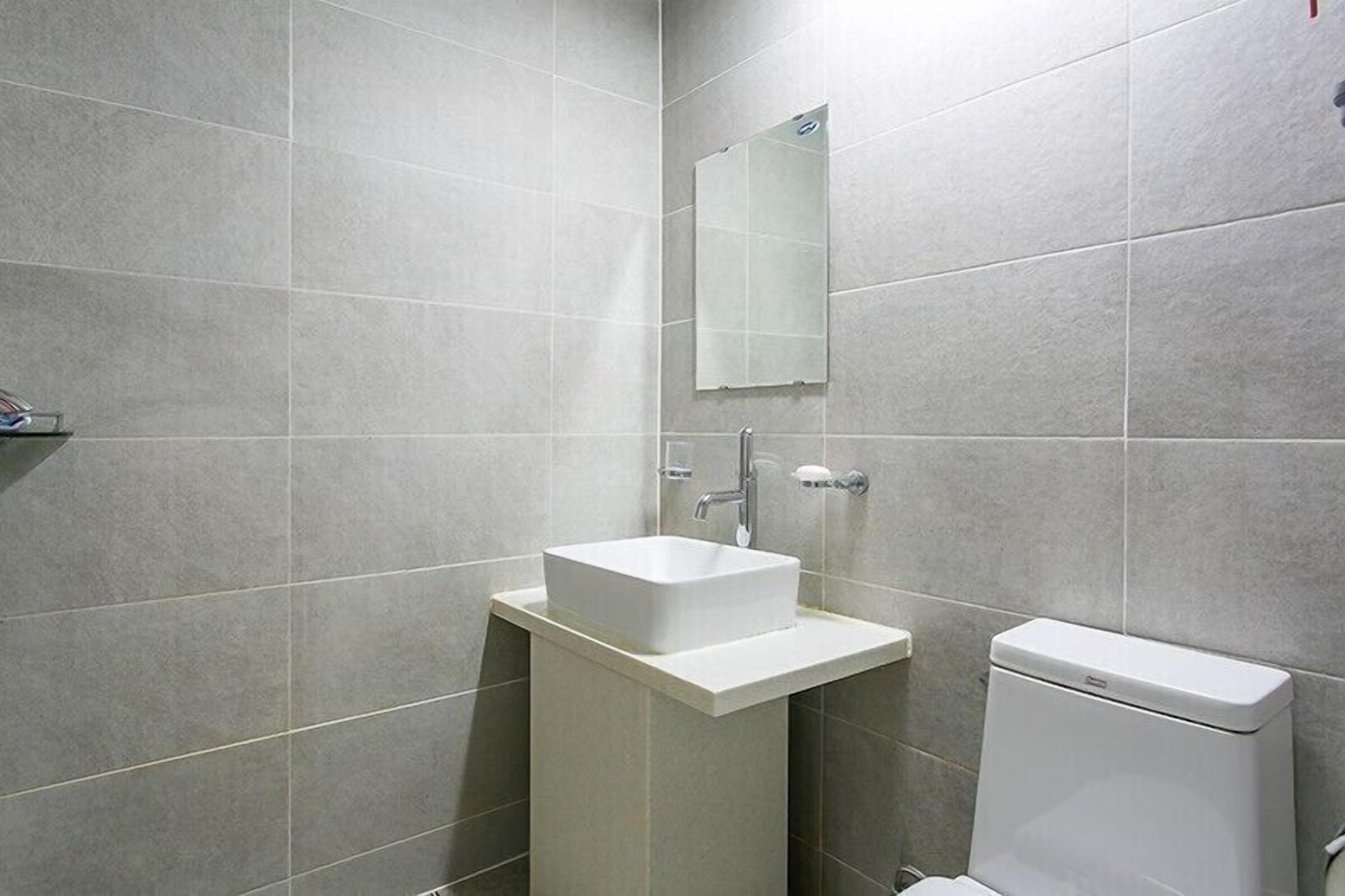 A bathroom with a white sink, mirror, and toilet against grey tiled walls.