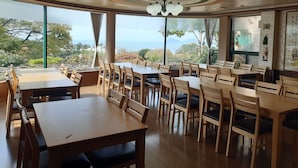Een restaurant met houten tafels en stoelen, grote ramen met uitzicht op zee, en een plafond met inbouwverlichting.