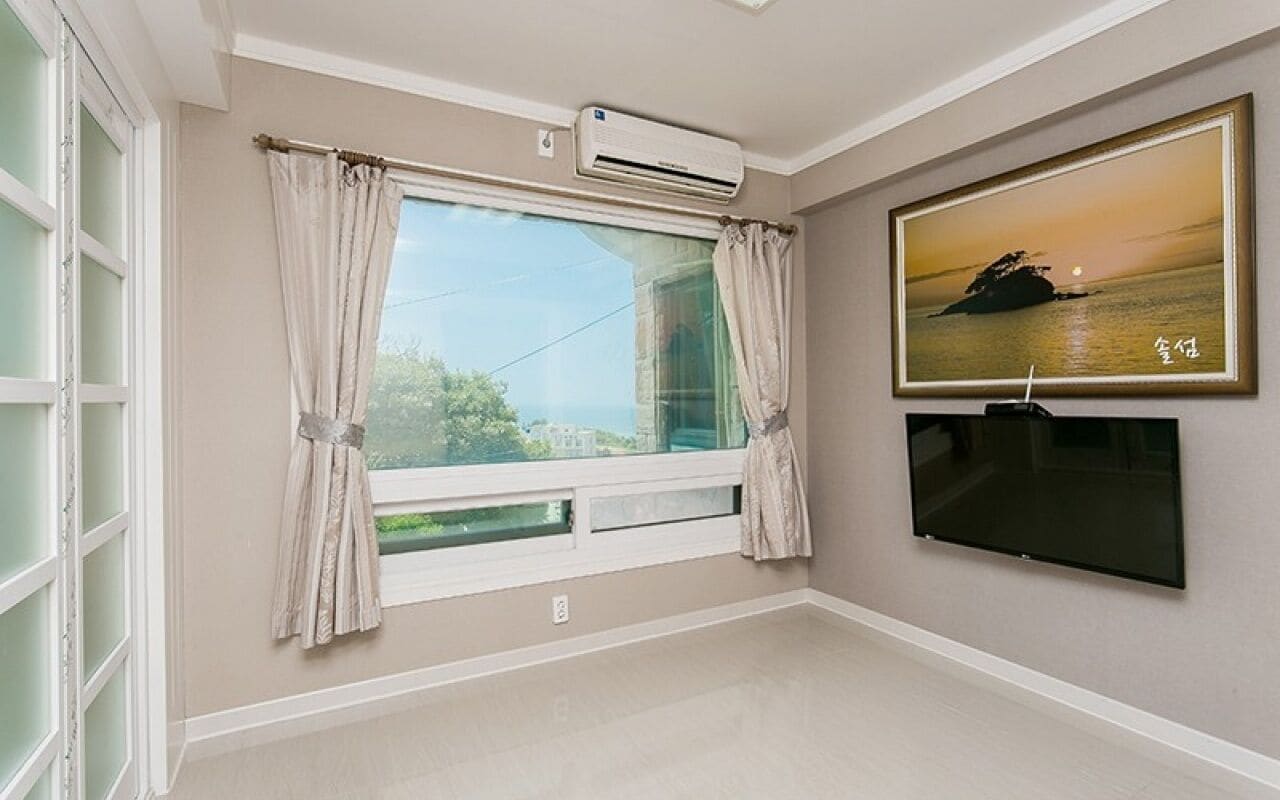 A room with a large window, an air conditioning unit, a flat-screen TV, and a framed picture on the wall.