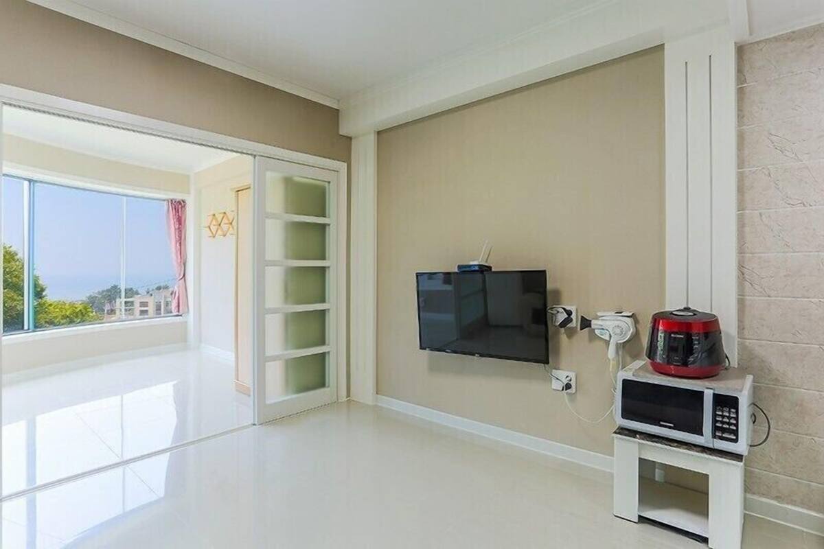 Una sala de estar moderna con un televisor de pantalla plana montado en la pared, un mueble blanco pequeño con un microondas y una puerta corrediza de vidrio que da a un balcón con vistas a la ciudad.
