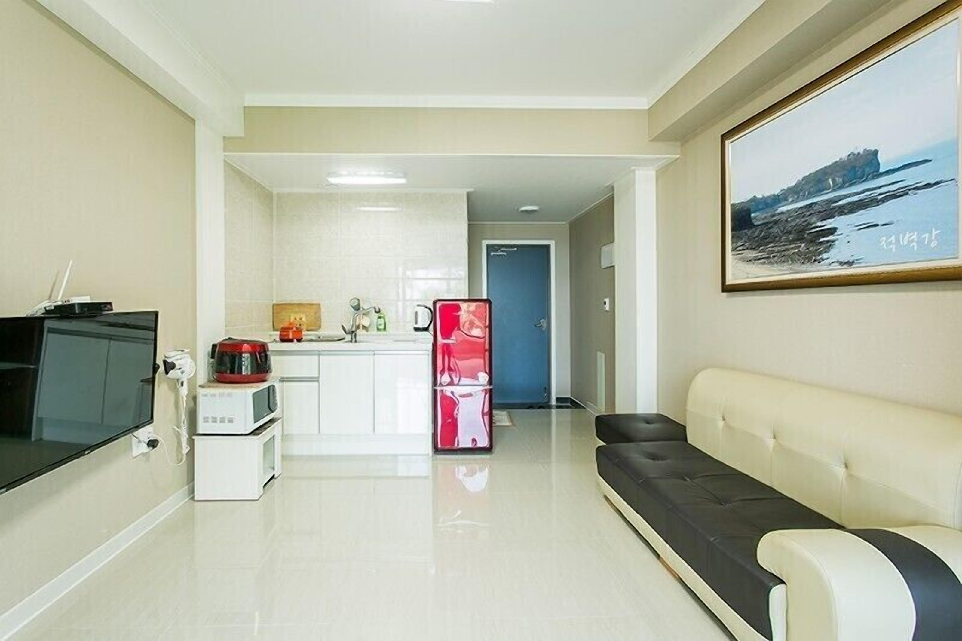 A modern living room with a sofa, a flat-screen TV, a kitchen area with a microwave, and a refrigerator.