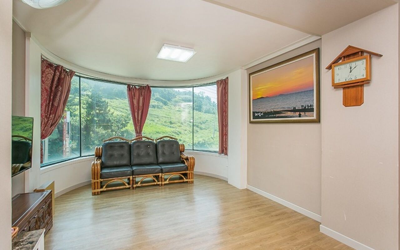 A room with a curved window, a wicker sofa, a clock, and a framed sunset painting.